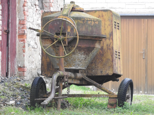 Alter rostiger Anhänger vor einem Backsteingebäude geparkt, umgeben von Gras und Steinen, mit einer Wand und einer Holztür im Hintergrund.