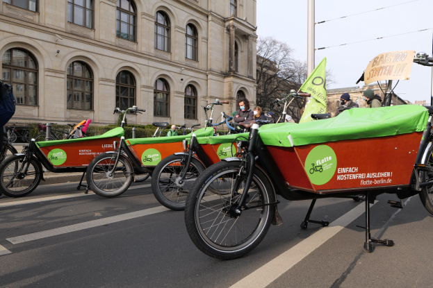 Eine Gruppe von Fahrrädern, die entlang einer Straße geparkt sind, mit einer Person in der Nähe, Gebäuden und Bäumen im Hintergrund unter einem klaren blauen Himmel, und einem Banner, das eine Fahrrad-Sharing-Kampagne anzeigt.