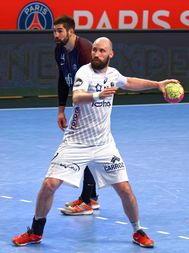 Zwei Männer spielen Handball auf einem Platz, einer hält den Ball, mit einer Tafel im Hintergrund, die 'Paris Saint-Germain vs Paris Saint Germain' anzeigt.