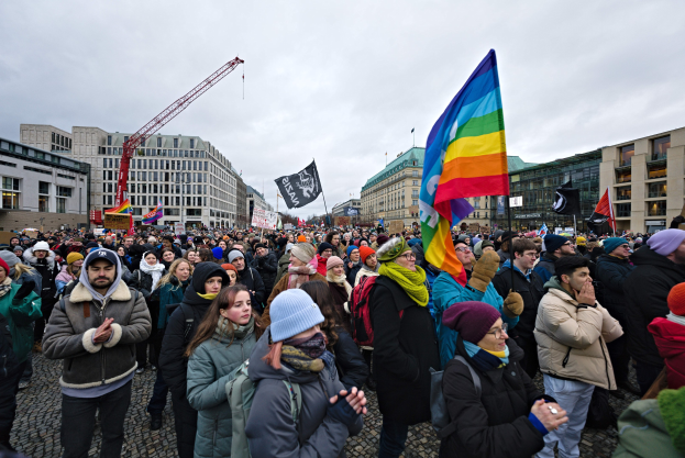Eine große Gruppe von Menschen hält LGBTQ+-Rechte-Schilder vor einem Gebäude mit einem Kran und einem bewölkten Himmel im Hintergrund.