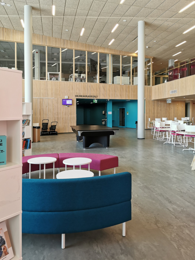Großer Raum mit mehreren Tischen, Stühlen, Sofas und Hockern in einer schulartigen Einrichtung, mit Bücherregalen, Deckenleuchten, Säulen und einem wandmontierten Fernseher.