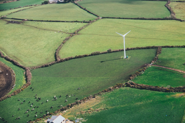 Luftaufnahme eines Windrades in einer grünen Wiese mit Bäumen, Häusern und Tieren, das sich in Irland befindet.