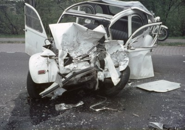 Ein schwer beschädigt aussehendes Auto mit einer zertrümten Front auf der Straße, mit Bäumen und einem Gebäude im Hintergrund und ein weiteres Fahrzeug in der Ferne.
