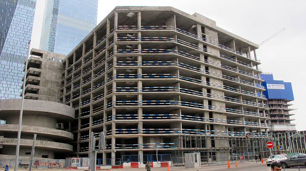 Großes Bauwerk im Bau in einer städtischen Umgebung mit Hochhäusern, Fahrzeugen, Fußgängern, Verkehrsinfrastruktur und Kränen vor einem klaren Himmel.