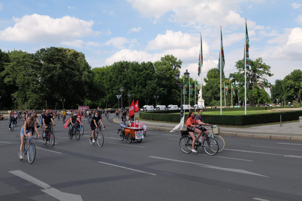 Eine Gruppe von Radfahrern fährt eine von Bäumen gesäumte Straße mit Laternenmasten und Fahnenmasten während der Pariser Radtour 2016, mit Fahrzeugen im Hintergrund unter einem bewölkten Himmel.