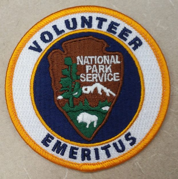 Rundes Abzeichen mit blauem Rand und weißem Hintergrund, mittig die schwarze Schrift "Volunteer National Park Service Emeritus" in einem gelben Kreis.