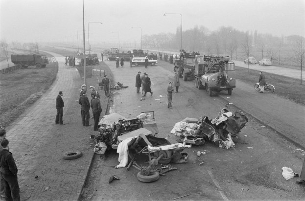 Schwarz-weißes Bild einer Autounfall-Szene an der Straße mit mehreren Fahrzeugen und einer Gruppe von Menschen drumherum, Lichtmasten, Bäume und Himmel im Hintergrund.
