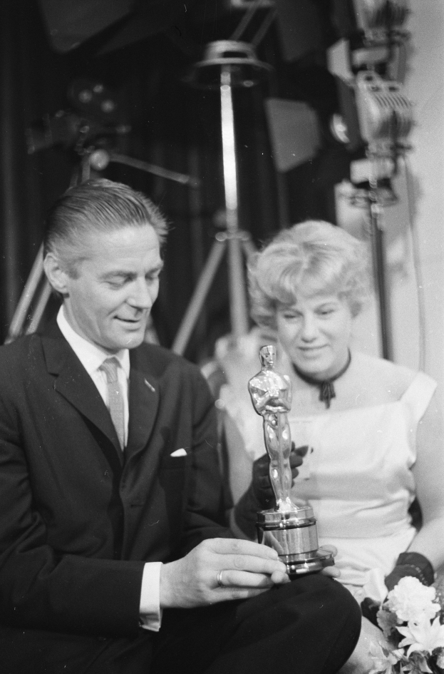 Ein Mann in einem schwarzen Blazer, weißem Hemd und Krawatte hält eine Oscar-Statue, während er neben einer Frau in Abendgarderobe sitzt, die einen Blumenstrauß hält, mit sanfter Beleuchtung im Hintergrund.