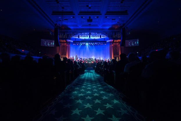 Großer Saal mit sitzendem Publikum, Bühne mit Musikern und Deckenbeleuchtung und Bildschirme, was auf einen Konzertsaal der Nationalen Convention 2019 hindeutet.