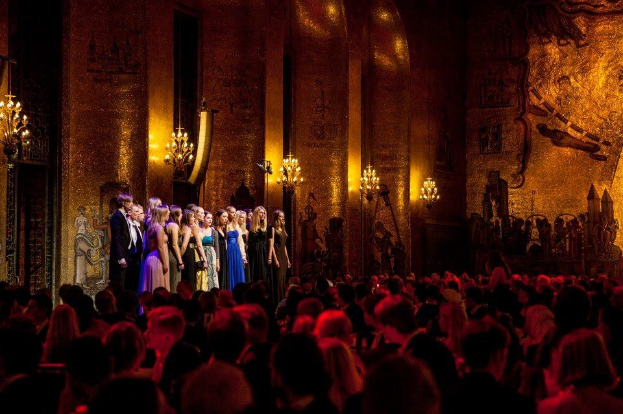 Eine Gruppe von Menschen auf einer beleuchteten Bühne mit bemalten Wänden im Royal Opera House, betrachtet von einem Publikum im Vordergrund.