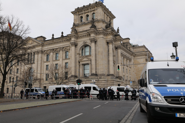 Eine Gruppe von Polizeibeamten steht vor dem Reichstagsgebäude in Berlin, Deutschland, mit Fahrzeugen, einem Zaun, Verkehrsampeln, Laternenpfählen, Bäumen und Fahnen im Hintergrund, bei klarem Himmel.