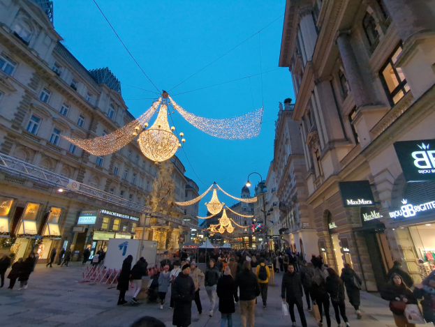 Eine belebte Stadtstraße voller Menschen, die unter festlichen Weihnachtslichtern an der Decke entlanggehen, mit Gebäuden auf beiden Seiten und einem klaren blauen Himmel im Hintergrund.