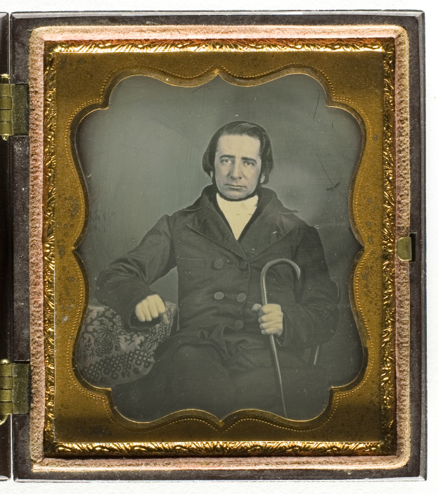 Ein altes Daguerreotyp eines Mannes in einem Anzug, der einen Stock hält, mit einem ernsten Gesichtsausdruck, der in die Ferne schaut.