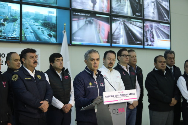 Eine Gruppe von Männern, die vor einem Podium mit einem Mikrofon und einer Tafel mit der Aufschrift 'Mexikanische Zoll- und Grenzschutzbehörde' stehen, mit Fernsehern und einer Flagge an der Wand dahinter.