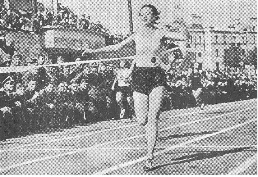 Ein Schwarz-Weiß-Foto einer Frau, die auf einer Laufbahn während des Frauen-100m-Finals bei den Olympischen Spielen 1956 läuft, mit einer Menschenmenge, Gebäuden und Pfählen im Hintergrund.