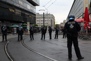 Polizeibeamte in schwarzer Uniform und Helm auf einer Straße mit Gebäuden, Glasfenstern und verstreuter Outdoor-Möbel, mit Bäumen und Strommasten im Hintergrund unter einem klaren Himmel.