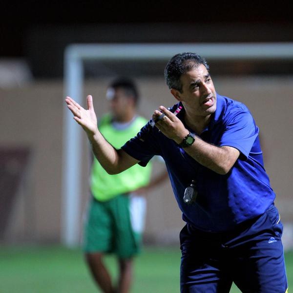 Ein Mann in einem blauen T-Shirt und Hose steht auf einem Fußballfeld und hält ein Mikrofon in der Hand, während er als Trainer gestikuliert. Im Hintergrund ist eine weitere Person in einem grünen Kleid, ein Torpfosten und eine Wand zu sehen.