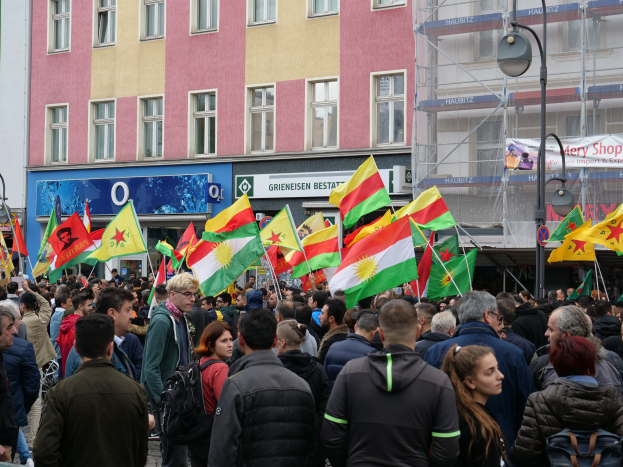 Eine große Gruppe von Menschen geht eine Straße entlang, hält kurdische Flaggen in den Händen, mit Straßenlaternen und Gebäuden am Straßenrand und Texttafeln im Hintergrund.
