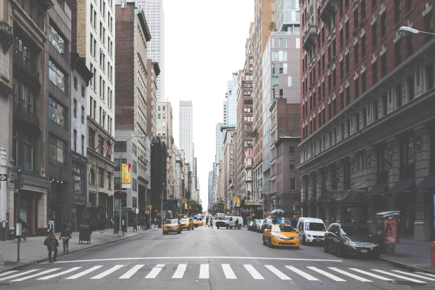 Eine belebte Stadtstraße mit hohen Gebäuden, Fahrzeugen, Fußgängern, Laternenmasten und Werbetafeln, die die Top-Attraktionen in New York City auflisten.