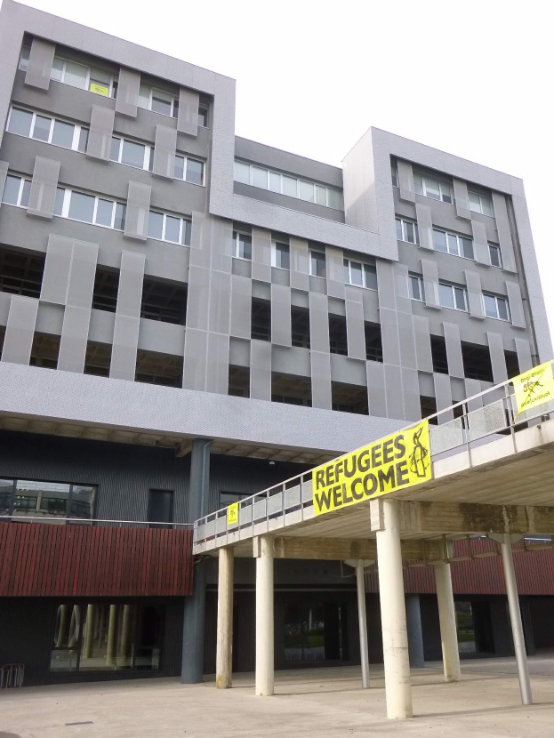 Gro├če Geb├Ąude mit einem "Flüchtlinge Willkommen"-Schild, umgeben von S├Ąulen und einem Gel├Ąnder, vor einem Himmel-Hintergrund.