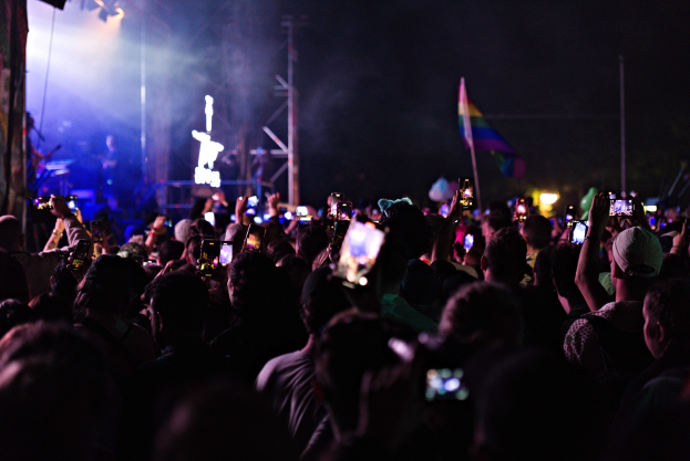 Eine Menge bei einem Konzert hält beleuchtete Handys hoch, während Menschen auf der Bühne unter Lichtern und Fahnen Instrumente spielen.