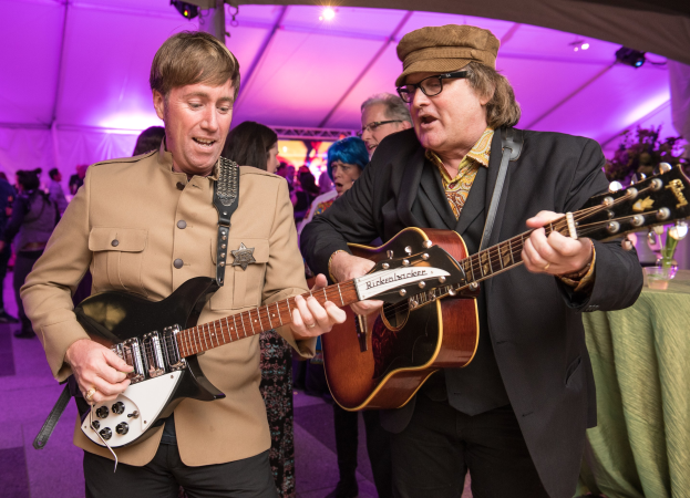 Zwei Männer spielen akustische Gitarren auf der Bühne bei der Induktionszeremonie des Rock and Roll Hall of Fame, vor einem Publikum und einem grünen Tisch mit Gläsern und einer Vase rechts, unter beleuchtetem Zeltdach.