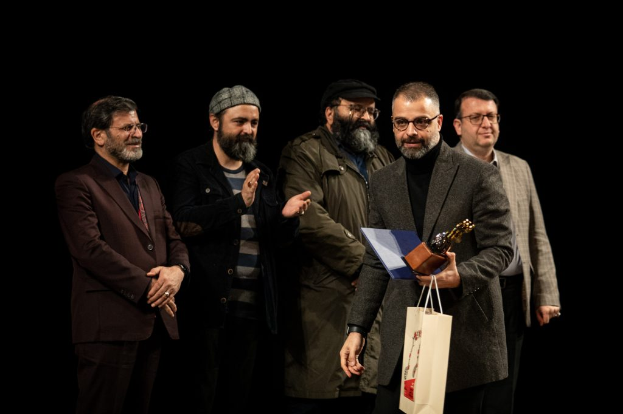 Eine Gruppe von Männern steht auf einer Bühne zusammen, einer hölt eine Tasche und eine Trophäe, mit einem dunklen Hintergrund, der die Eröffnung der Berliner Filmfestspiele suggeriert.