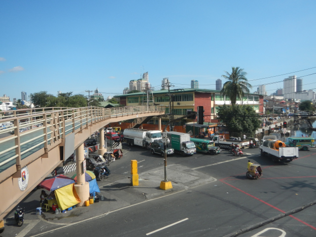 Eine belebte Straße mit Schwerverkehr neben einer Brðcke, mit Fahrzeugen, Fðßlern, Zelten, Versorgungsmästen, Ampeln, Bäumen, Gebäuden und einer Wasserstraße im Hintergrund bei einem bewölktem Himmel.