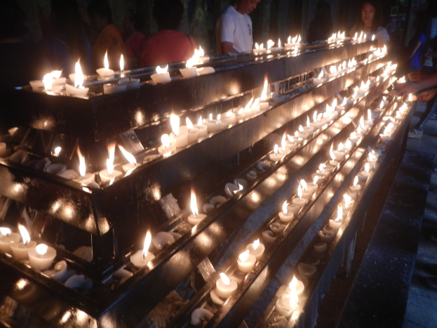 Eine Gruppe von Menschen steht um eine lange Reihe von Kerzen in einer Kirche, beleuchtet von ihrem flackernden Licht gegen eine Wand im Hintergrund.