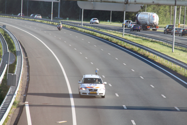 Ein Auto auf einer Autobahn mit einem stationären Polizeiwagen am Straßenrand, andere Fahrzeuge im Verkehr und Bäume und Straßenstrukturen im Hintergrund.
