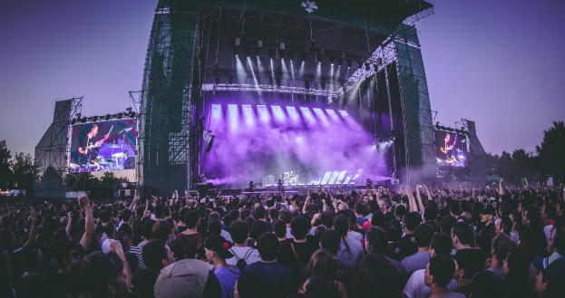 Eine große Menschenmenge auf einem Musikfestival schaut Performern auf einer hell erleuchteten Bühne mit Instrumenten und Bildschirmen zu, die vor einem Gebäude, Bäumen und einem klaren blauen Himmel aufgebaut ist.