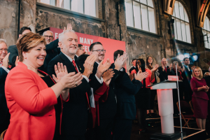Eine Gruppe von Menschen, die vor einem Publikum applaudieren, mit einem Podium, einem Mikrofon und einer Tafel mit Text auf der rechten Seite und Stühlen, einer Fahne, einer Wand, Fenstern und Lichtern im Hintergrund.