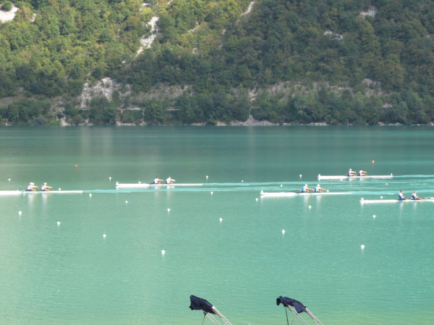 Gruppe von Menschen bei einem Ruderwettbewerb auf einem Gewässer mit Bäumen und einem Hügel im Hintergrund, Kleidung über Stühle drapiert.
