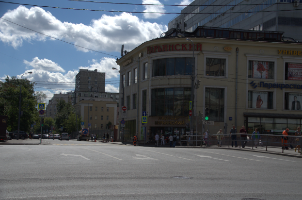 Eine Stadtstraße mit Menschen, hohen Gebäuden, Schildern, Ampeln, einem Metallzaun, Fahrzeugen, Bäumen und einem bewölkten Himmel.