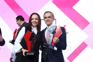 Zwei Personen auf einer Bühne mit Trophäen stehend und strahlend, mit einem Awards-Ceremony-Hintergrund.