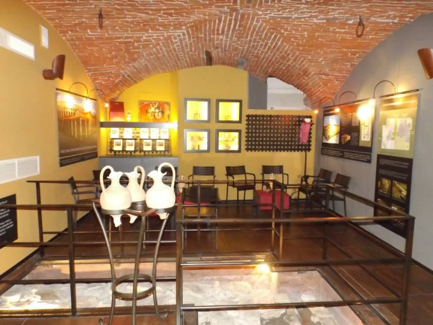 Ein Museumsraum mit mehreren Tischen, Stühlen, Displaytafeln, gerahmten Kunstwerken an den Wänden, Vasen, Leuchten und anderen Gegenständen.