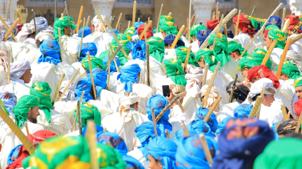 Eine große Gruppe von Menschen in bunten Kostümen marschiert auf einer Straße, jeder hält einen Stock in der Hand, mit einem Gebäude im Hintergrund während eines Festivals.