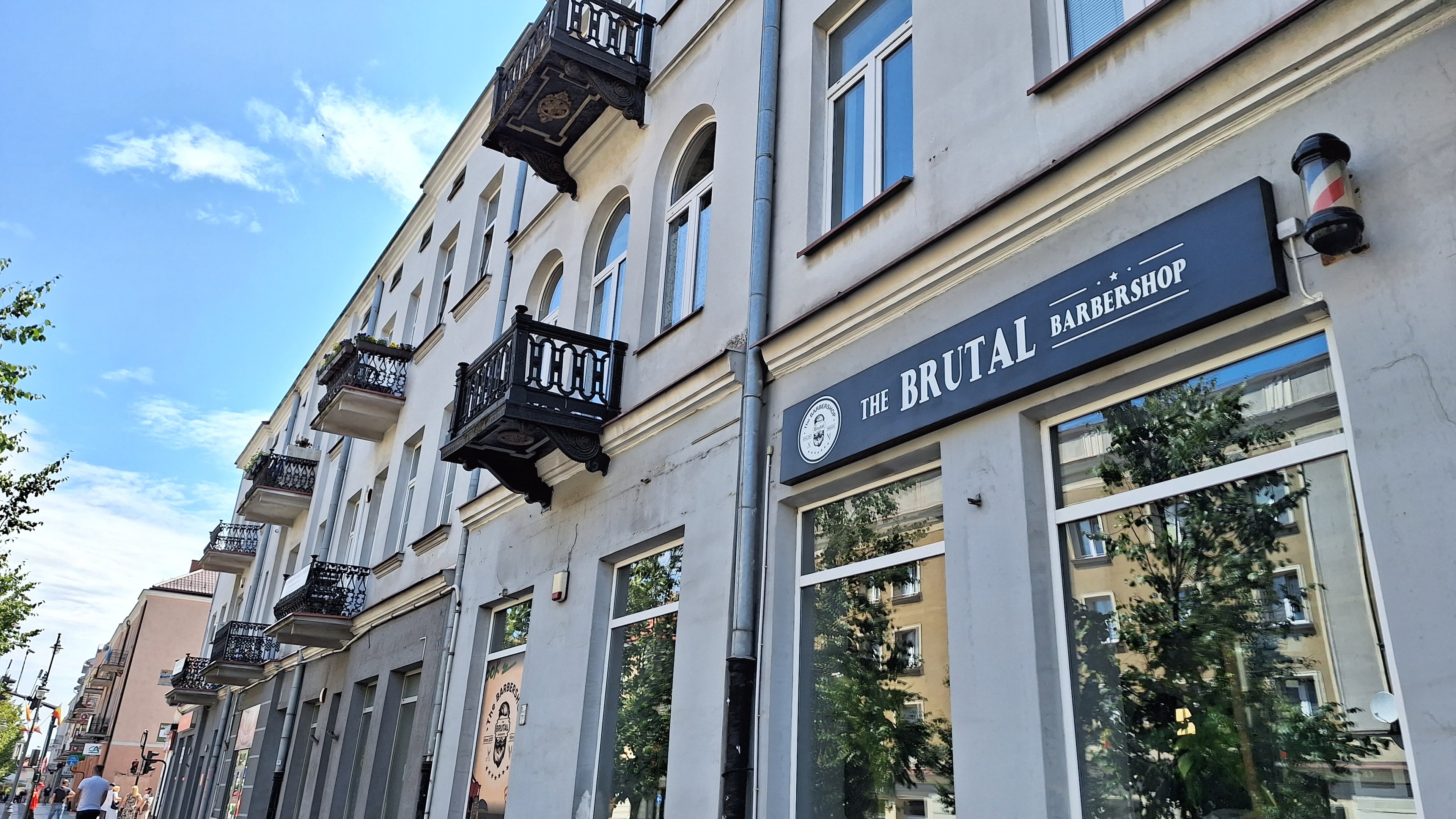 Brutalistische Barbershop-Fassaden in Berlin mit Betongebäuden, Fenstern, Geländern, Balkonen und Schildern, umgeben von Bäumen, Straßenlaternen, Fußgängern und einer bewölkten Himmel