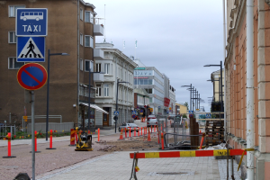 Stadtstraße mit Gebäuden, Straßeninfrastruktur, Fahrzeugen, Bäumen und einer Baustelle mit Verkehrsschildern unter einem bewölkten Himmel.