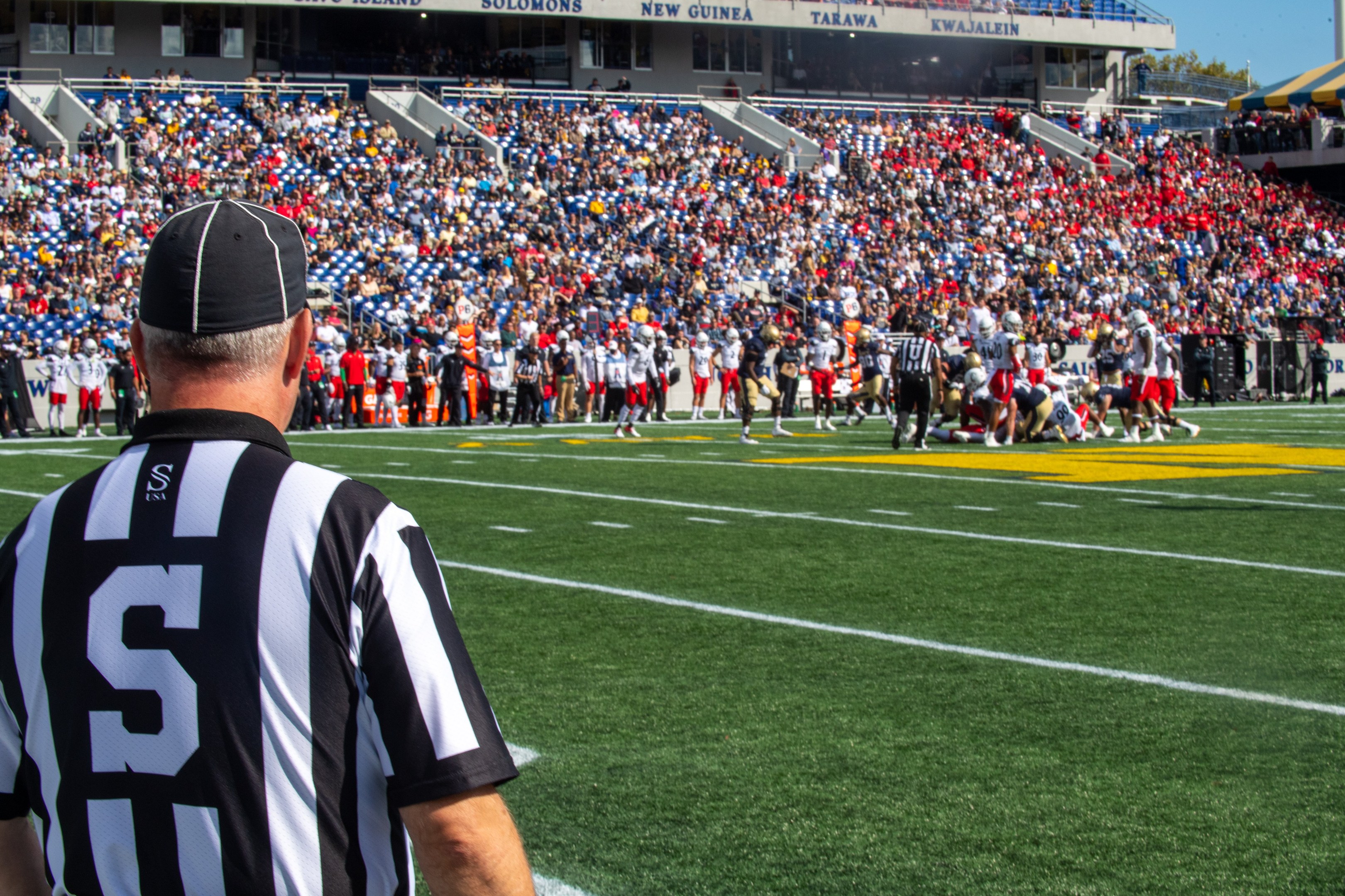 Referee standing on a football field beside spectators, some wearing caps, with railings, boards, poles, trees, and a clear blue sky in the background.