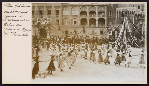 Ein Schwarz-Weiß-Foto von Menschen in traditioneller Kleidung, die vor einem Gebäude mit Fenstern, Bögen, Säulen und Geländern tanzen, mit einem Pfahl mit Gegenständen rechts und Text links, das einen Karneval in Paris vermuten lässt.