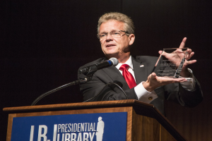 Ein Mann im Anzug und Krawatte hält eine Auszeichnung auf einem Podium mit Mikrofon und Tafel, vor einem dunklen Hintergrund bei einer Bibliothek-Veranstaltung.