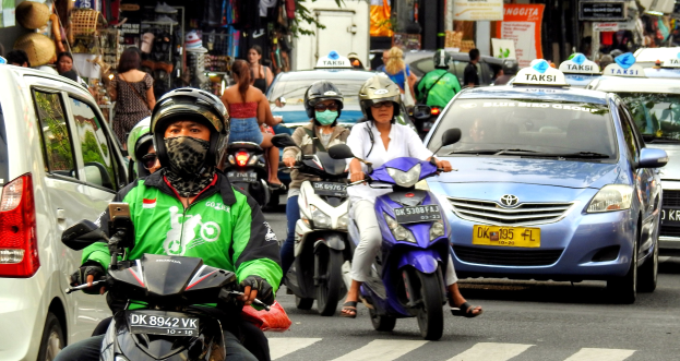 Eine Gruppe von Menschen, die auf Motorradfahrern die Straße entlangfahren, mit Autos und anderen Fahrzeugen, einige Fahrer tragen Helme, mit Gebäuden, Bäumen, Schildern mit Text und Geschäften im Hintergrund.