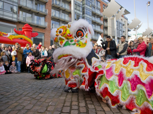 Ein lebendiges chinesisches Neujahrsfest in Amsterdam mit einem Löwen tanzen im Vordergrund mit einer Menschenmenge, einige halten Kameras, die sich um es versammelt, vor einem Hintergrund von Gebäuden, Laternenmasten und einem klaren blauen Himmel.
