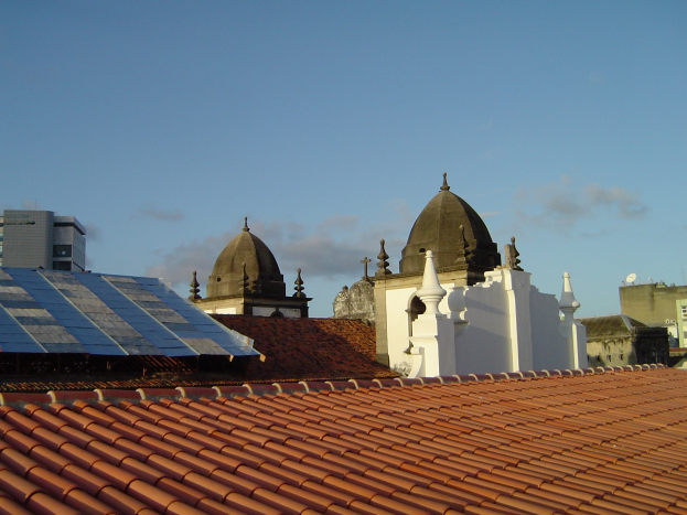 Stadtpanorama mit Gebäuden im Vordergrund, Solarmodule auf einem Dach und einem blauen Himmel im Hintergrund.