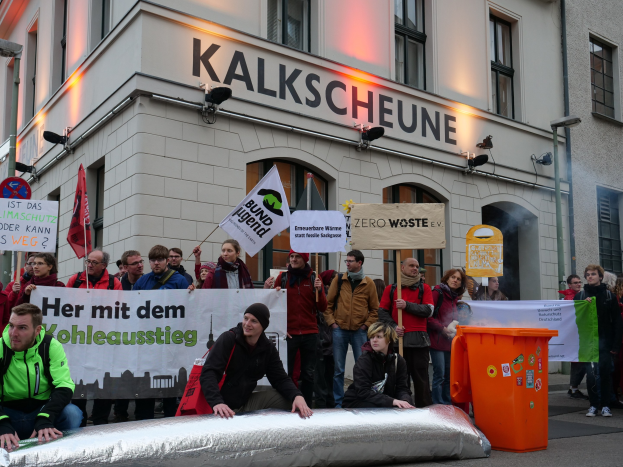 Eine Gruppe von Menschen mit Schildern und Transparenten vor einem Gebäude, zwei Personen im Vordergrund und ein Müllcontainer rechts, während einer Protestaktion in Deutschland.