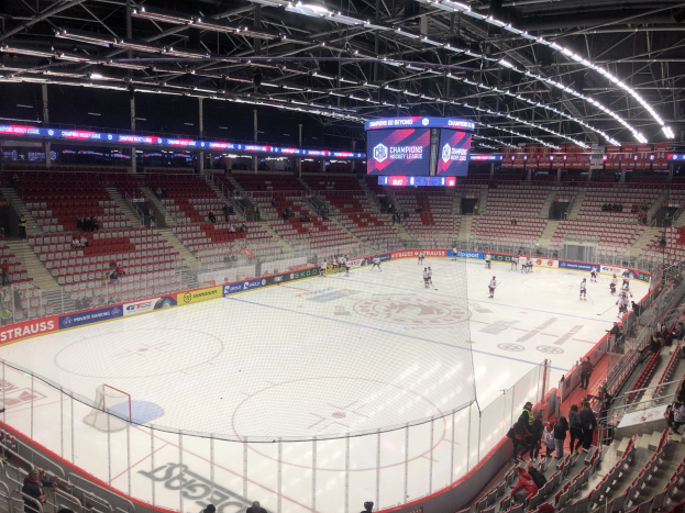 Eishockeyspiel in einer großen Arena mit Zuschauern, ein Rink umgeben von einem Zaun, ein Display, das "St. Louis Blues vs Detroit Red Wings" anzeigt, Metallrahmen, ein Dach mit Deckenleuchten und ein Dach.
