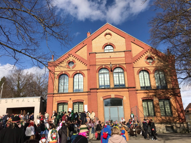 Eine große Gruppe von Menschen steht vor einem mehrstöckigen Gebäude mit Fenstern und Türen im Freien, umgeben von Bäumen unter einem bewölkten Himmel, möglicherweise bei einem Stadtfest.