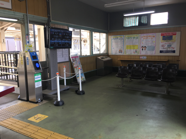Ein belebter Bahnhofseingang mit Reihen von Stühlen, einem Servicecounter, Ticketautomaten, Deckenleuchten, Geländern, Informationsanzeigen und Wandplakaten.