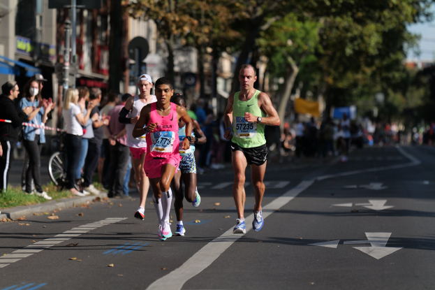 Gruppe von Menschen, die bei einem Stadtmarathon laufen, mit Zuschauern auf der linken Seite, unscharfen städtischen Hintergrund mit Bäumen, Gebäuden und einem Fahrrad.